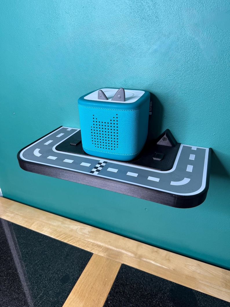May include: A teal speaker with a perforated design is on a road-themed shelf. The shelf is black and grey with white road markings and a chequered flag. The speaker has grey ear-shaped accents on top.