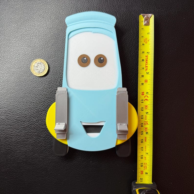 May include: A light blue, yellow, gray, and black 3D-printed Mater car-shaped phone holder. The holder is approximately 8 inches tall. The design includes Mater's eyes, mouth, and tow hooks. A yellow measuring tape is next to the holder.