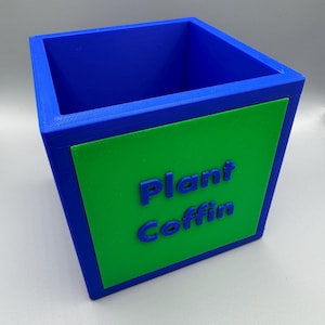 May include: A blue and green rectangular planter with the words "Plant Coffin" in blue. The planter is a cube shape with a green rectangle on the front. The interior is blue.