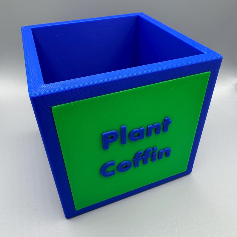 May include: A blue and green rectangular planter with the words "Plant Coffin" in blue. The planter is a cube shape with a green rectangle on the front. The interior is blue.