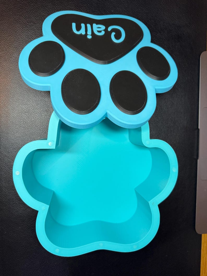 Paw print-shaped pet memorial box with personalised name on the lid. Holds up to 1300ml, ideal for pet ashes or keepsakes. Customisable colours available. Secure magnetic closure. Thoughtful gift for dog or cat owners. Designed for home display.