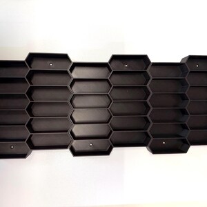 May include: A black wall-mounted storage organizer featuring a repeating honeycomb pattern with multiple hexagonal compartments. The structure is composed of interconnected cells arranged in a horizontal grid, with small circular mounting holes visible.