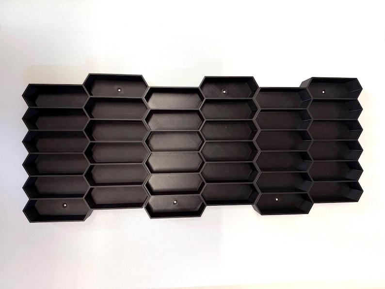 May include: A black wall-mounted storage organizer featuring a repeating honeycomb pattern with multiple hexagonal compartments. The structure is composed of interconnected cells arranged in a horizontal grid, with small circular mounting holes visible.