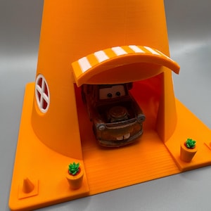 Op de afbeelding: Een oranje speelhuis in de vorm van een verkeerskegel met een wit afdak en een kleine speelgoedauto erin. Het speelhuis heeft twee kleine groene potplanten aan de zijkanten.