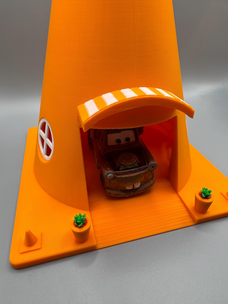 Op de afbeelding: Een oranje speelhuis in de vorm van een verkeerskegel met een wit afdak en een kleine speelgoedauto erin. Het speelhuis heeft twee kleine groene potplanten aan de zijkanten.