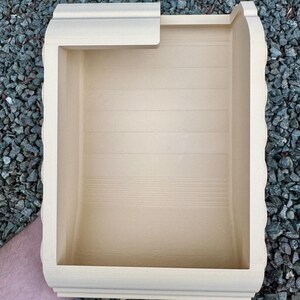 May include: A light beige, rectangular, open-top container with a smooth, matte finish. The interior features horizontal recessed lines along the back wall, and the top edge has a partial overhang on one side, creating a structured, hollow form.