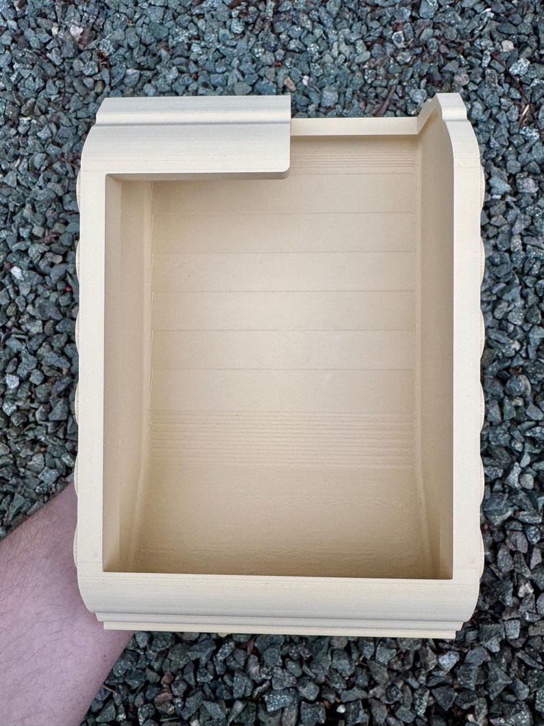 May include: A light beige, rectangular, open-top container with a smooth, matte finish. The interior features horizontal recessed lines along the back wall, and the top edge has a partial overhang on one side, creating a structured, hollow form.