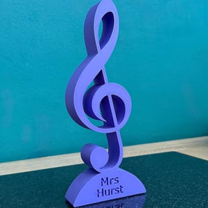 K&ouml;nnte beinhalten: Ein violettes, 3D-gedrucktes Notensymbol in Form eines Violinschl&uuml;ssels, das aufrecht auf einem geschwungenen Sockel steht. Auf dem Sockel ist der Text Mrs Hurst in einer klaren, serifenlosen Schrift eingraviert. Das Objekt ist matt.