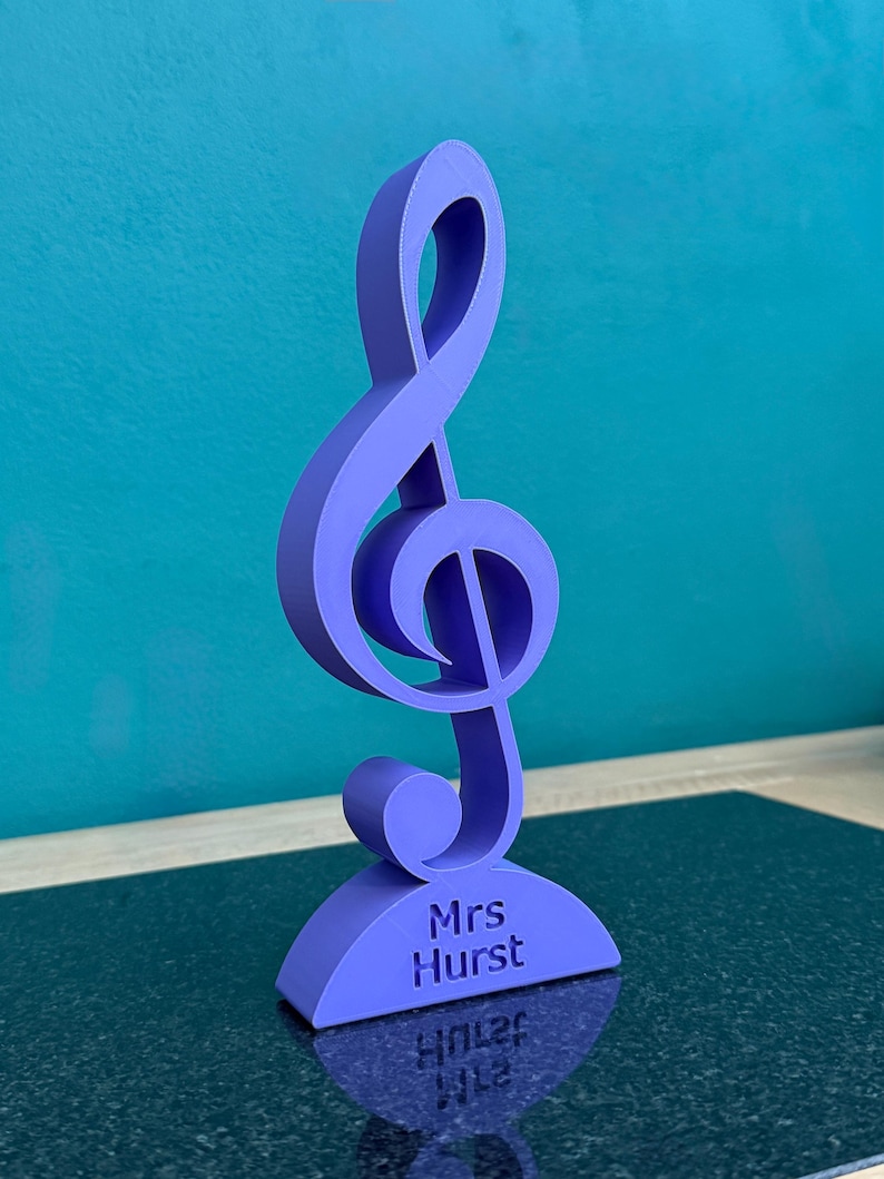K&ouml;nnte beinhalten: Ein violettes, 3D-gedrucktes Notensymbol in Form eines Violinschl&uuml;ssels, das aufrecht auf einem geschwungenen Sockel steht. Auf dem Sockel ist der Text Mrs Hurst in einer klaren, serifenlosen Schrift eingraviert. Das Objekt ist matt.