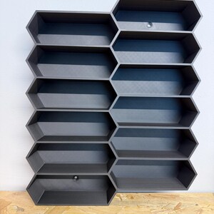 May include: A dark gray wall-mounted storage unit featuring a modular honeycomb design with ten hexagonal compartments arranged in two vertical columns. The structure is made of a matte material and includes two visible circular mounting holes.