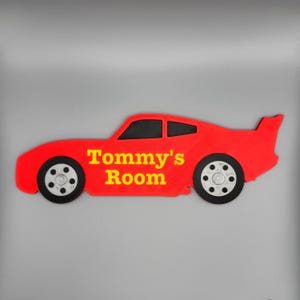 May include: A red car-shaped sign with the text "Tommy's Room" in yellow. The sign has black wheels and windows. The car has a spoiler on the back.