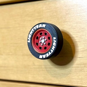 May include: A black and red drawer knob shaped like a tire, with the word "LIGHTYEAR" in white lettering around the tire. The center of the tire is red with a silver hubcap. The knob is designed to look like a car wheel.