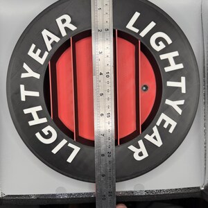 May include: A black and red circular object with the text "LIGHTYEAR" in white. The object has a series of red vertical bars in the center. A ruler is placed on the object, measuring approximately 12 inches.
