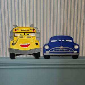 May include: A yellow school bus character wall decoration with a grey bumper, a chain detail, and the text SCHOOL BUS, positioned next to a blue car character wall decoration with white headlights and a grey grille, both featuring expressive eyes.
