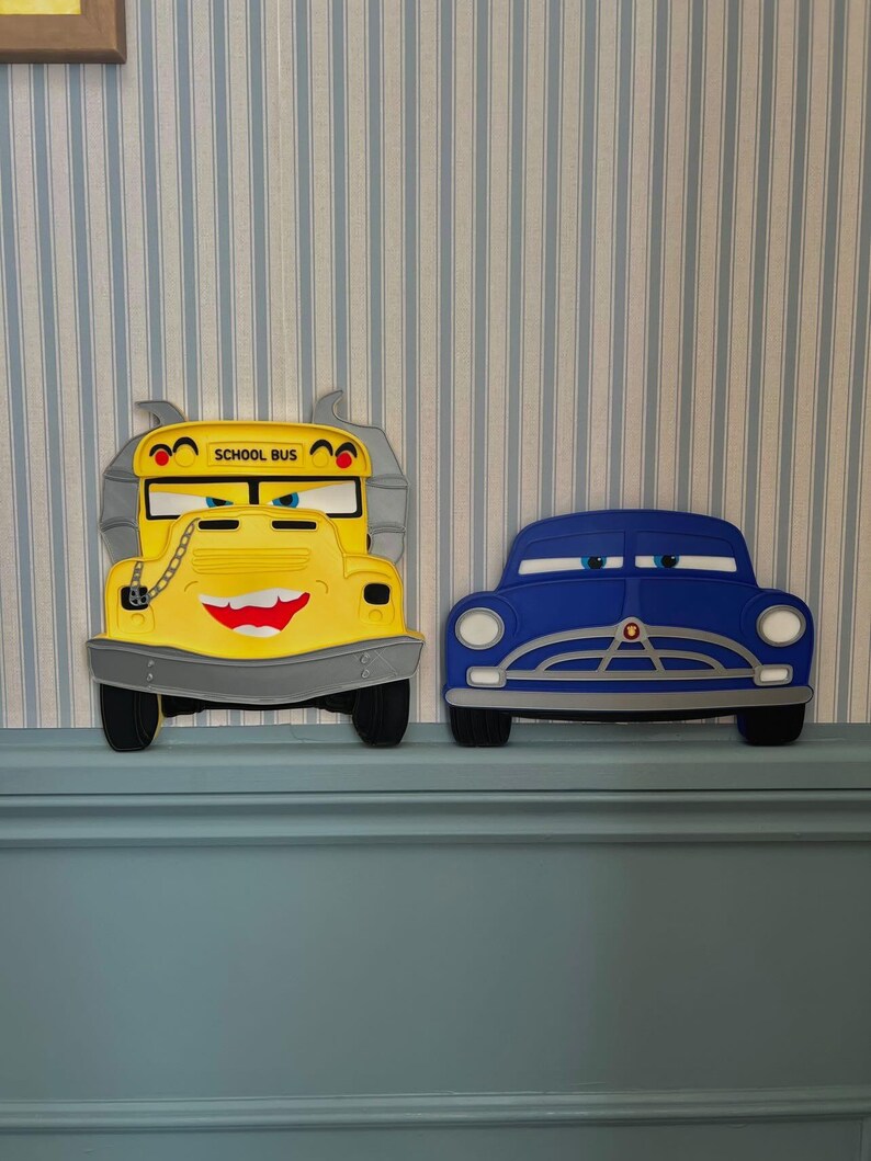 May include: A yellow school bus character wall decoration with a grey bumper, a chain detail, and the text SCHOOL BUS, positioned next to a blue car character wall decoration with white headlights and a grey grille, both featuring expressive eyes.