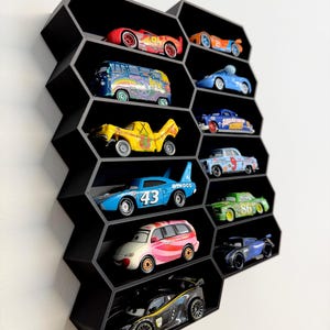 May include: A black honeycomb-shaped wall display shelf with multiple hexagonal compartments. Each compartment holds a miniature die-cast toy car, including various colorful vehicles with unique designs, racing numbers, and character-inspired graphics.