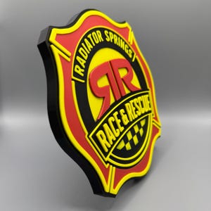 May include: A shield-shaped emblem with the words "RADIATOR SPRINGS RACE & RESCUE" in a yellow and black design. The emblem has a red and yellow border with the letters "RR" in red. The background is a light gray.