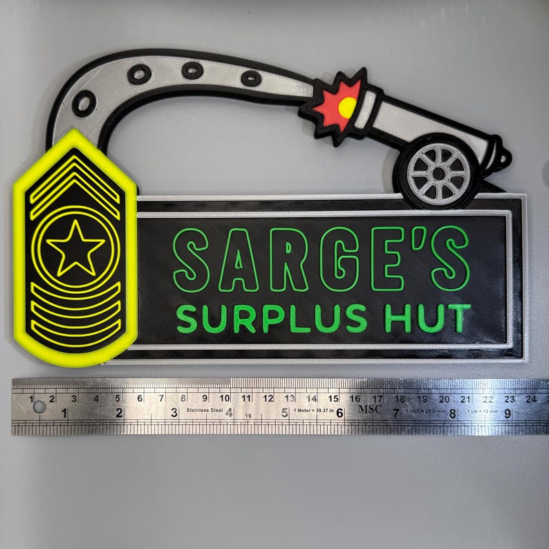 May include: A decorative sign featuring a silver cannon and a yellow and black military rank insignia. The sign reads "SARGE'S SURPLUS HUT" in green lettering on a black background. A ruler shows the sign is approximately 9 inches long.