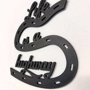May include: Black metal wall art in the shape of a horseshoe with the words "Life is a highway" in a cursive font. The art piece is mounted on a white wall. The horseshoe design has a textured surface.
