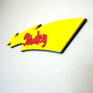 May include: A yellow sign with the name "Stanky" in red script. The sign has a black border and is mounted on a white wall. The sign is a decorative element.