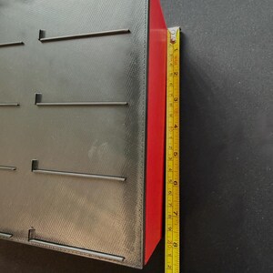 May include: A black rectangular object with slots and a red side is measured with a yellow tape measure. The object is approximately 8 inches tall. The tape measure is green and yellow.