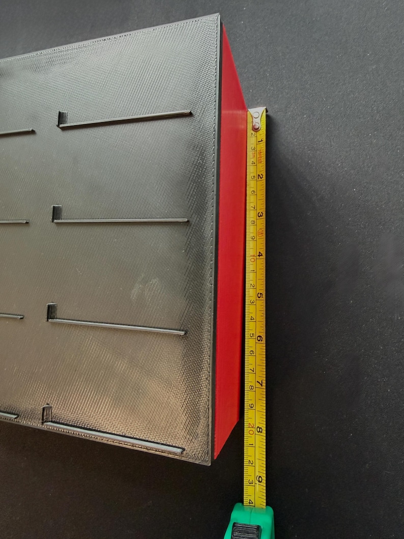 May include: A black rectangular object with slots and a red side is measured with a yellow tape measure. The object is approximately 8 inches tall. The tape measure is green and yellow.