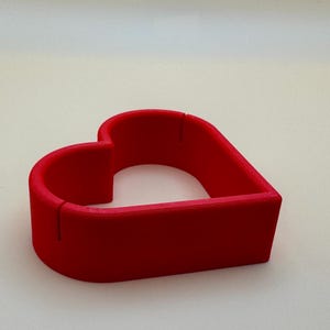 A red 3D-printed heart-shaped place card holder, perfect for weddings and romantic events. these holders can be customised in your choice of colours to match your theme, making them ideal for unique table décor and personalised celebrations.