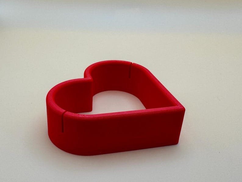 A red 3D-printed heart-shaped place card holder, perfect for weddings and romantic events. these holders can be customised in your choice of colours to match your theme, making them ideal for unique table décor and personalised celebrations.