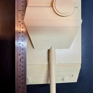 May include: A cream-colored, unpainted plastic model tank turret with a long barrel and a circular hatch. A stainless steel ruler is positioned along the left side, showing the model measures approximately 8.5 inches in total length.