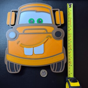 May include: An orange and brown 3D-printed Mater from the Cars movie. The character has white teeth, green eyes, and a smiling face. The item is approximately 19 inches tall, as measured by a yellow tape measure.