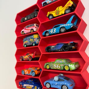 May include: A bright red wall-mounted display shelf with a honeycomb pattern, featuring ten individual hexagonal compartments. Each compartment holds a single die-cast toy car, including various colorful models with racing numbers and graphic designs.