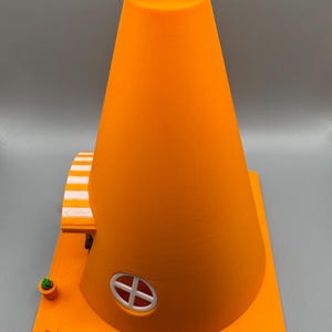 Op de afbeelding: Een 3D-geprint oranje verkeerskegelhuis met een wit gestreept luifel, een klein raam en een witte antenne op de top.