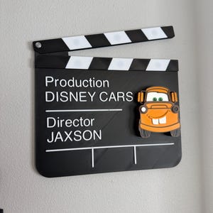 May include: A black and white film clapperboard with the text "Production DISNEY CARS Director JAXSON". An orange cartoon car is attached to the clapperboard. The clapperboard is open.