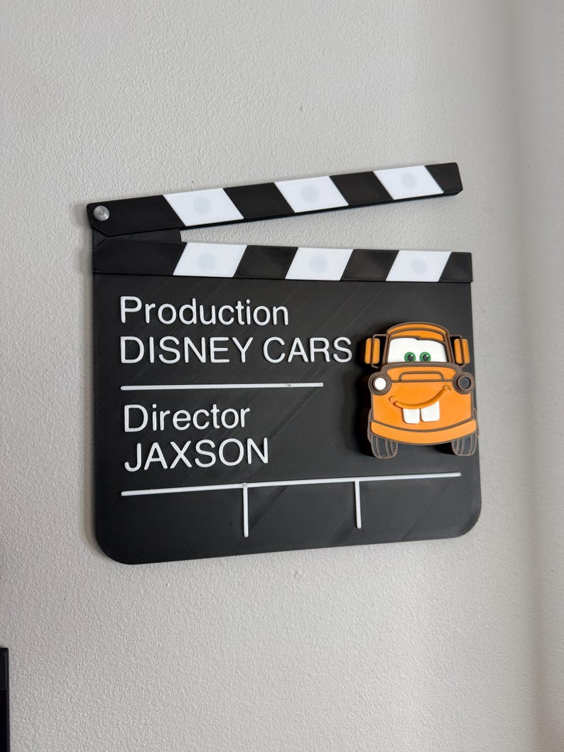 May include: A black and white film clapperboard with the text "Production DISNEY CARS Director JAXSON". An orange cartoon car is attached to the clapperboard. The clapperboard is open.