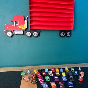 May include: A red wall-mounted display shelf shaped like a semi-truck with a trailer featuring multiple horizontal slots for small toy cars. The truck cab has a yellow lightning bolt graphic and the number 95. Numerous small toy cars are arranged below.