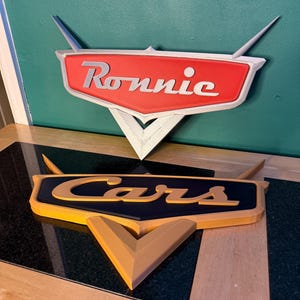 May include: Two 3D-printed signs, one reading "Ronnie" in white on a red background, and the other reading "Cars" in yellow on a black background. Both signs have a stylized design with a triangular base and wing-like extensions.