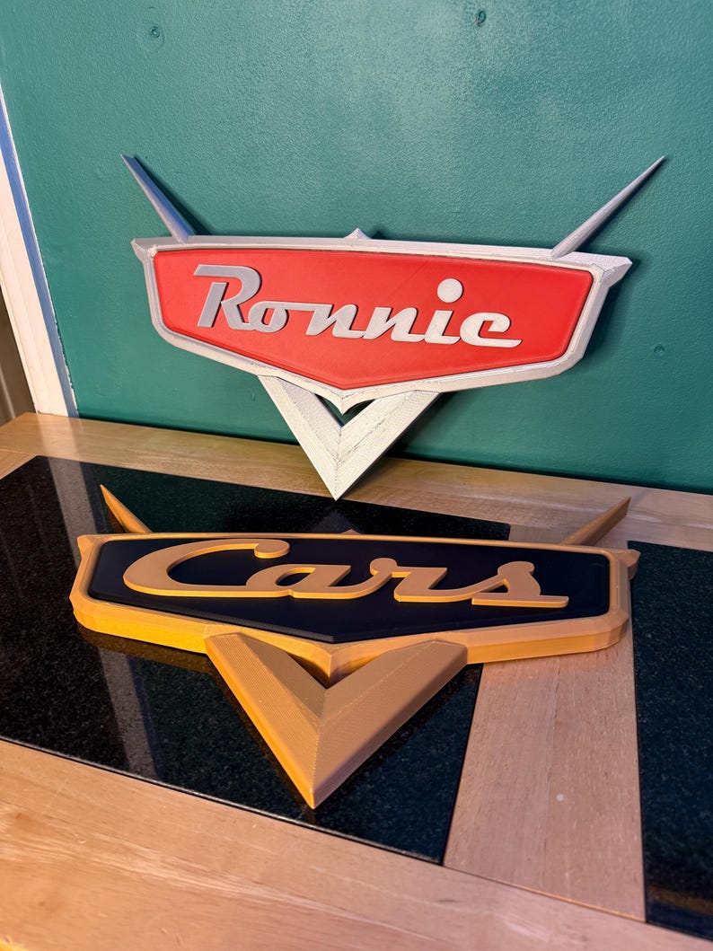 May include: Two 3D-printed signs, one reading "Ronnie" in white on a red background, and the other reading "Cars" in yellow on a black background. Both signs have a stylized design with a triangular base and wing-like extensions.