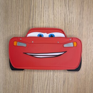K&ouml;nnte beinhalten: Ein rotes Lightning McQueen-f&ouml;rmiges Accessoire mit einem l&auml;chelnden Gesicht, blauen Augen und wei&szlig;en Akzenten. Das Accessoire hat schwarze Reifen und ein detailliertes Design, perfekt f&uuml;r Fans des Animationsfilms Cars.