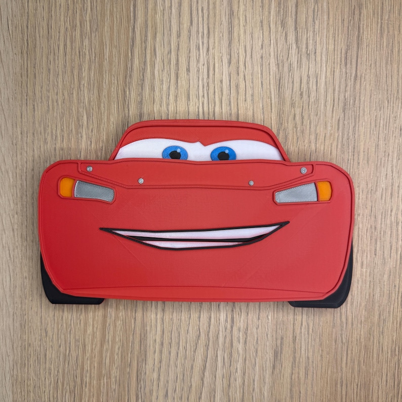 K&ouml;nnte beinhalten: Ein rotes Lightning McQueen-f&ouml;rmiges Accessoire mit einem l&auml;chelnden Gesicht, blauen Augen und wei&szlig;en Akzenten. Das Accessoire hat schwarze Reifen und ein detailliertes Design, perfekt f&uuml;r Fans des Animationsfilms Cars.