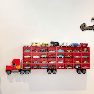 May include: A red truck-shaped shelf filled with various toy cars. The shelf is mounted on a white wall and features a detailed design of a truck with wheels and a trailer. The toy cars are of different colors and styles.