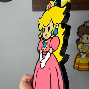 Op de afbeelding: Een 3D-geprinte Princess Peach-figuur uit de Super Mario-videogameserie. De figuur heeft een roze jurk, geel haar en een blauwe kroon. De figuur is zwart omlijnd en heeft een cartoonachtig ontwerp.