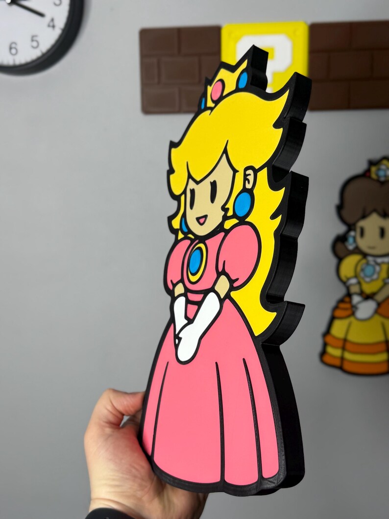 Op de afbeelding: Een 3D-geprinte Princess Peach-figuur uit de Super Mario-videogameserie. De figuur heeft een roze jurk, geel haar en een blauwe kroon. De figuur is zwart omlijnd en heeft een cartoonachtig ontwerp.