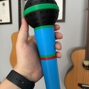 A 3D-printed, colorful plastic microphone prop held in a hand. This vibrant and durable design is perfect for photo booth props, party accessories, or as a fun kids' toy, available in various color combinations.