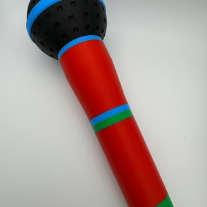 A 3D-printed, colorful plastic microphone prop held in a hand. This vibrant and durable design is perfect for photo booth props, party accessories, or as a fun kids' toy, available in various color combinations.