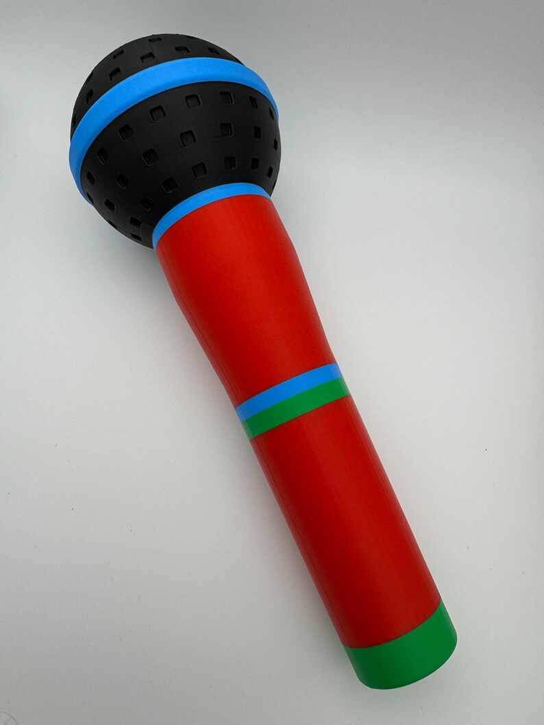 A 3D-printed, colorful plastic microphone prop held in a hand. This vibrant and durable design is perfect for photo booth props, party accessories, or as a fun kids' toy, available in various color combinations.