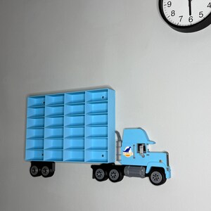 May include: A light blue toy truck-shaped wall shelf with multiple compartments. The truck has black wheels and a dinosaur logo on the side. A black and white clock is visible in the background.