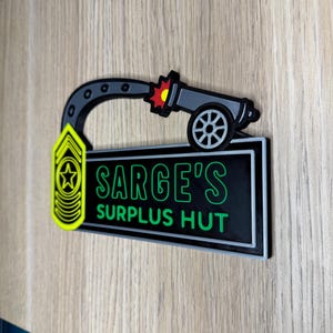 May include: A black and green sign with the text "SARGE'S SURPLUS HUT" in green lettering. The sign features a cartoon cannon firing a red and yellow burst. A yellow and black military insignia is on the left.