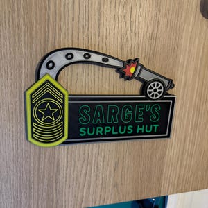 May include: A 3D-printed sign featuring a cartoon cannon firing a projectile. The sign includes the text "SARGE'S SURPLUS HUT" in green and white, and a yellow and black military insignia. The design is mounted on a wooden surface.
