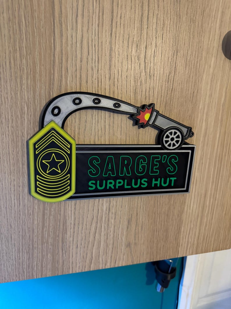May include: A 3D-printed sign featuring a cartoon cannon firing a projectile. The sign includes the text "SARGE'S SURPLUS HUT" in green and white, and a yellow and black military insignia. The design is mounted on a wooden surface.