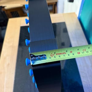 May include: A black, curved object with blue knobs along the edge. A yellow and green tape measure shows a width of approximately 2 inches. The object appears to be made of metal.